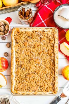 an apple crumble pie on a white table surrounded by apples, cinnamons and nuts