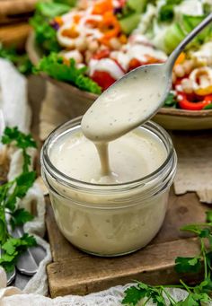 a spoon is pouring dressing into a glass bowl with salad in the background and fresh vegetables on the side