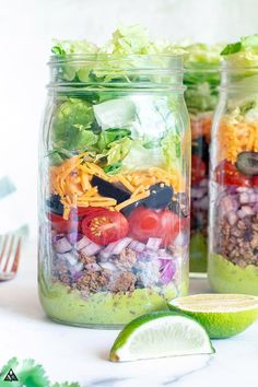 two mason jars filled with taco salad and topped with lettuce, tomatoes, black olives, avocado