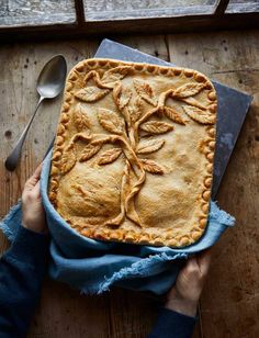 a person holding a pie on top of a wooden table next to a knife and fork