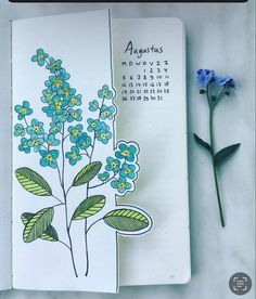 an open notebook with blue flowers on it next to a flower marker and some green leaves