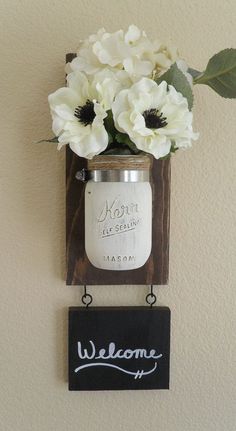 a mason jar with flowers in it hanging on a wall next to a welcome sign