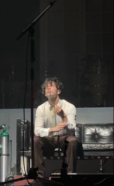 a man sitting in front of a microphone on top of a stage wearing a white shirt and brown pants