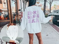 a woman standing on the sidewalk with her back turned to the camera, wearing a white sweatshirt that says i'm my dance era