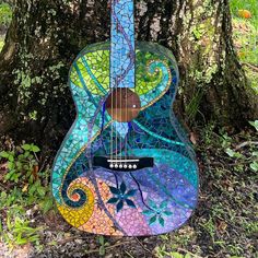 a colorful guitar sitting in the grass next to a tree with lots of leaves on it