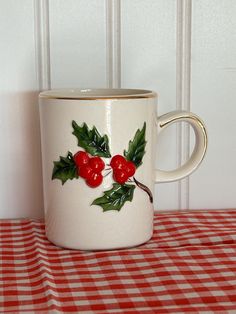 a white coffee mug with holly and red berries on it sitting on a checkered tablecloth