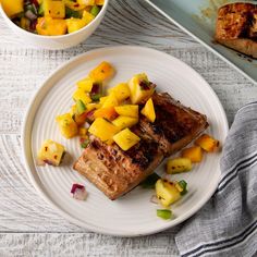 a white plate topped with fish and pineapple salsa next to a bowl of fruit