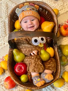 a baby sitting in a basket filled with apples, pears and oranges while wearing a thanksgiving hat