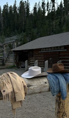 a cowboy hat, blanket and jacket on a fence post in front of a log cabin