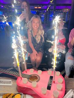 a woman sitting in front of a pink guitar cake with sparklers on it and people standing around