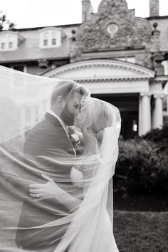 a bride and groom kissing in front of a large house with a veil blowing in the wind