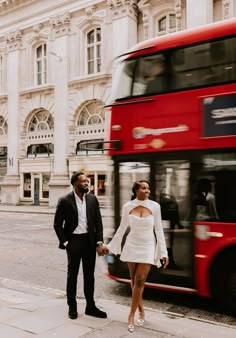 a man and woman standing in front of a red double decker bus on the street