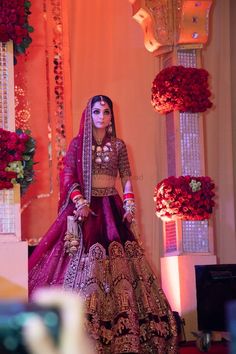 a woman in a purple and gold bridal outfit standing on a stage with flowers behind her