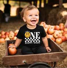 a baby sitting in a wooden crate with apples on the ground and one happy dude t - shirt