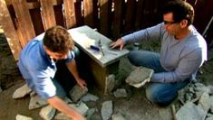 two men are working on some rocks in the back yard, and one man is using a hammer