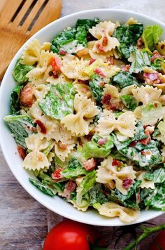 a white bowl filled with pasta salad next to tomatoes and lettuce on a wooden table