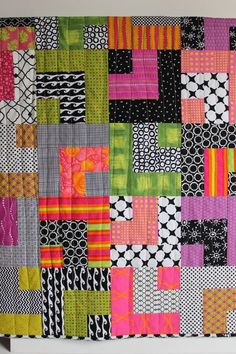 a colorful patchwork quilt hanging on the wall in front of a white frame with black, pink, green and orange squares