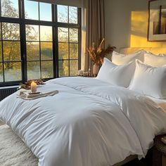 a bed with white comforters and pillows in front of a large window that overlooks the trees outside