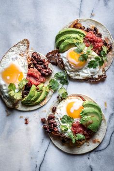 three tacos with eggs, meat and avocado on them sitting on a marble surface