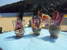 three glasses filled with sand sitting on top of a blue cloth covered table next to the ocean