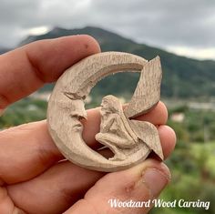 a hand holding a carved wooden brooch depicting a woman sitting on the moon with mountains in the background