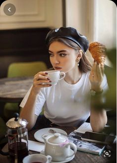 a woman sitting at a table with a cup and saucer in her hand while holding a pastry