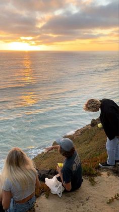 three people are sitting on the edge of a cliff overlooking the ocean at sunset or sunrise