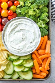 a tray filled with veggies and dip surrounded by tomatoes, broccoli, cucumbers, carrots, and chips