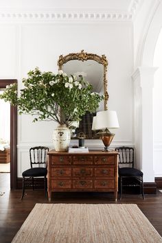 a wooden dresser topped with flowers next to a mirror and two chairs in front of it