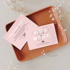 two pink business cards sitting on top of a brown tray next to white baby's breath flowers