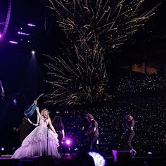 lady in white dress performing on stage with other people around her and fireworks behind her