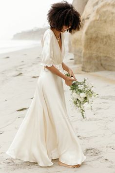 a woman standing on top of a sandy beach next to the ocean holding a bouquet