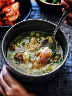 a person holding a spoon in a bowl of soup with meat and vegetables on the side