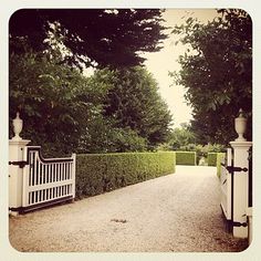 a white gate is in the middle of a path between two hedges and trees on either side