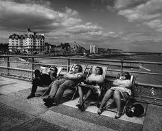 black and white photograph of four people sitting on a bench near the water with buildings in the background