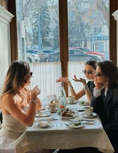 three women sitting at a table with food and drinks in front of them, talking