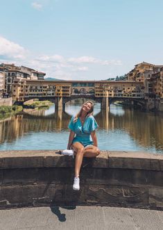 a woman sitting on the edge of a wall next to a river with buildings in the background