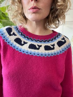 a woman wearing a pink sweater with blue and white designs on the front, standing in front of a potted plant