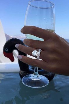 a hand holding a wine glass with a diamond ring on it, in front of a swan