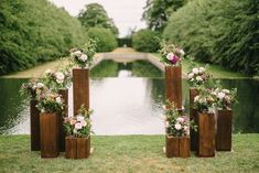 four tall wooden vases with flowers in them on the grass near some water and trees