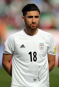 a man with a beard standing in front of a soccer field wearing a white shirt