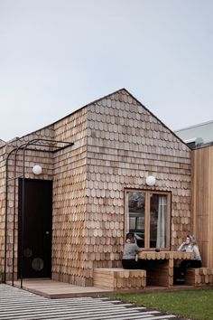 two people sitting on wooden benches in front of a building with shingled roofing