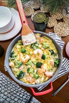 a skillet filled with pasta, shrimp and broccoli on top of a wooden table