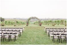 an outdoor ceremony set up with white folding chairs and floral arch over the aisle, surrounded by greenery