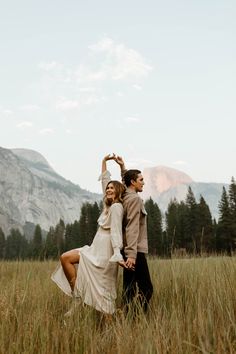 a man and woman standing in tall grass with mountains in the background