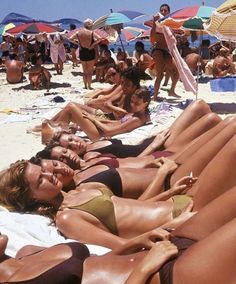 many people are laying on the beach in bikinis and sunbathers with umbrellas