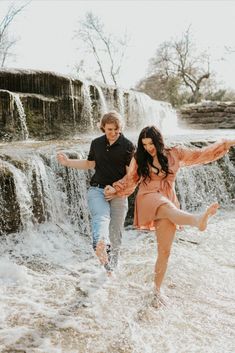 a man and woman are running through the water in front of a waterfall with their arms outstretched