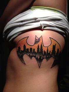 a batman tattoo on the side of a woman's stomach, with an orange and black city skyline behind it