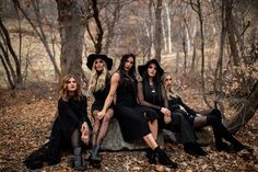 four women sitting on a rock in the woods, all wearing black dresses and hats