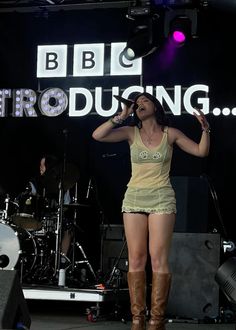 a woman in short shorts and boots singing into a microphone at an outdoor concert on stage